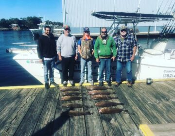 Group of happy anglers posing with their fresh grouper catch on a wooden dock in Crystal River, FL, with a charter fishing boat in the background.