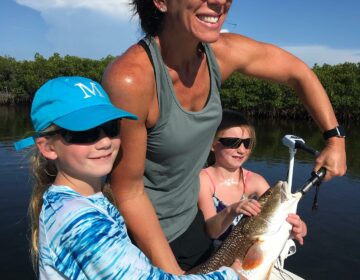 Smiling woman and two young girls on a boat holding a large redfish, dressed in sun-protective clothing and sunglasses, with water and greenery in the background.