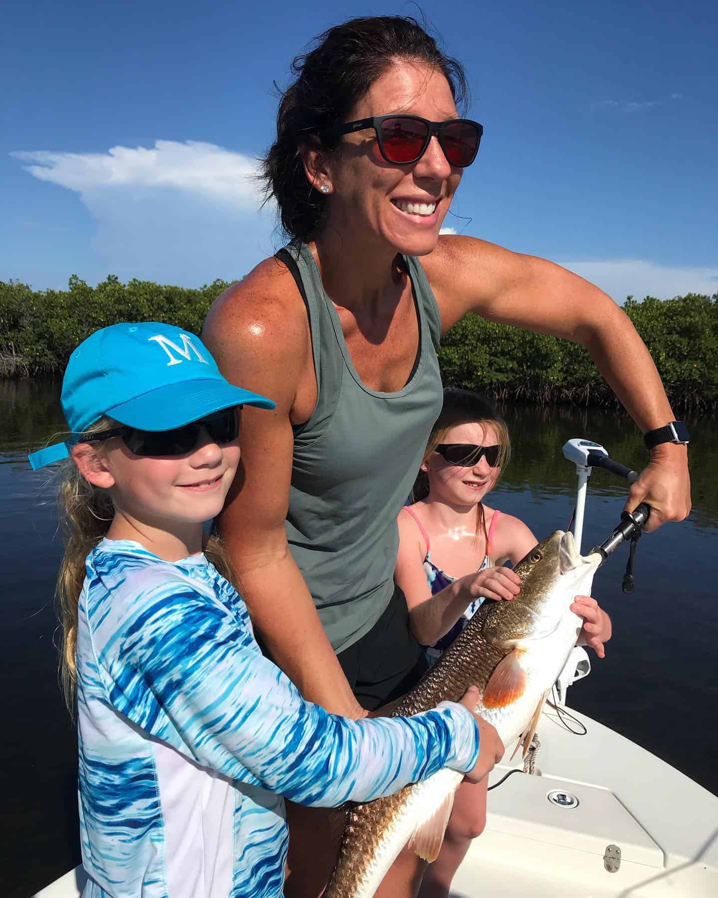 Smiling woman and two young girls on a boat holding a large redfish, dressed in sun-protective clothing and sunglasses, with water and greenery in the background.