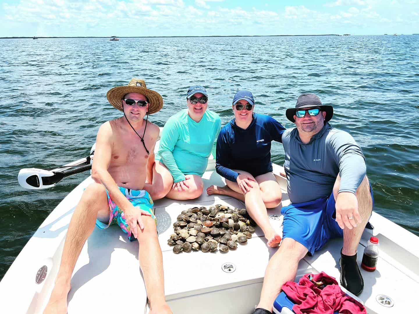 Four people on a boat with a pile of scallops, dressed in sun gear, smiling against a backdrop of open water and a bright sky.